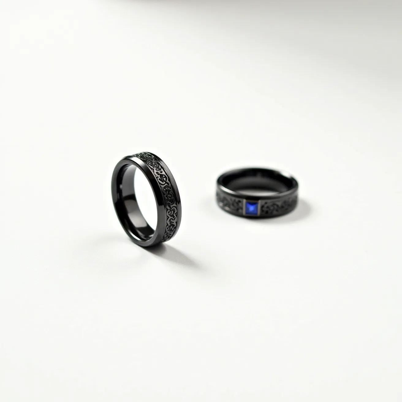 These tungsten rings feature a black finish with an intricate Celtic pattern engraved along the outer band. One of the rings is set with a square-cut blue gemstone, likely a sapphire or similar stone, secured in a bezel setting that complements the design. The bands are smooth and polished, enhancing their modern and stylish appearance, and they lack any clasps or additional attachments, showcasing a sleek, continuous design.