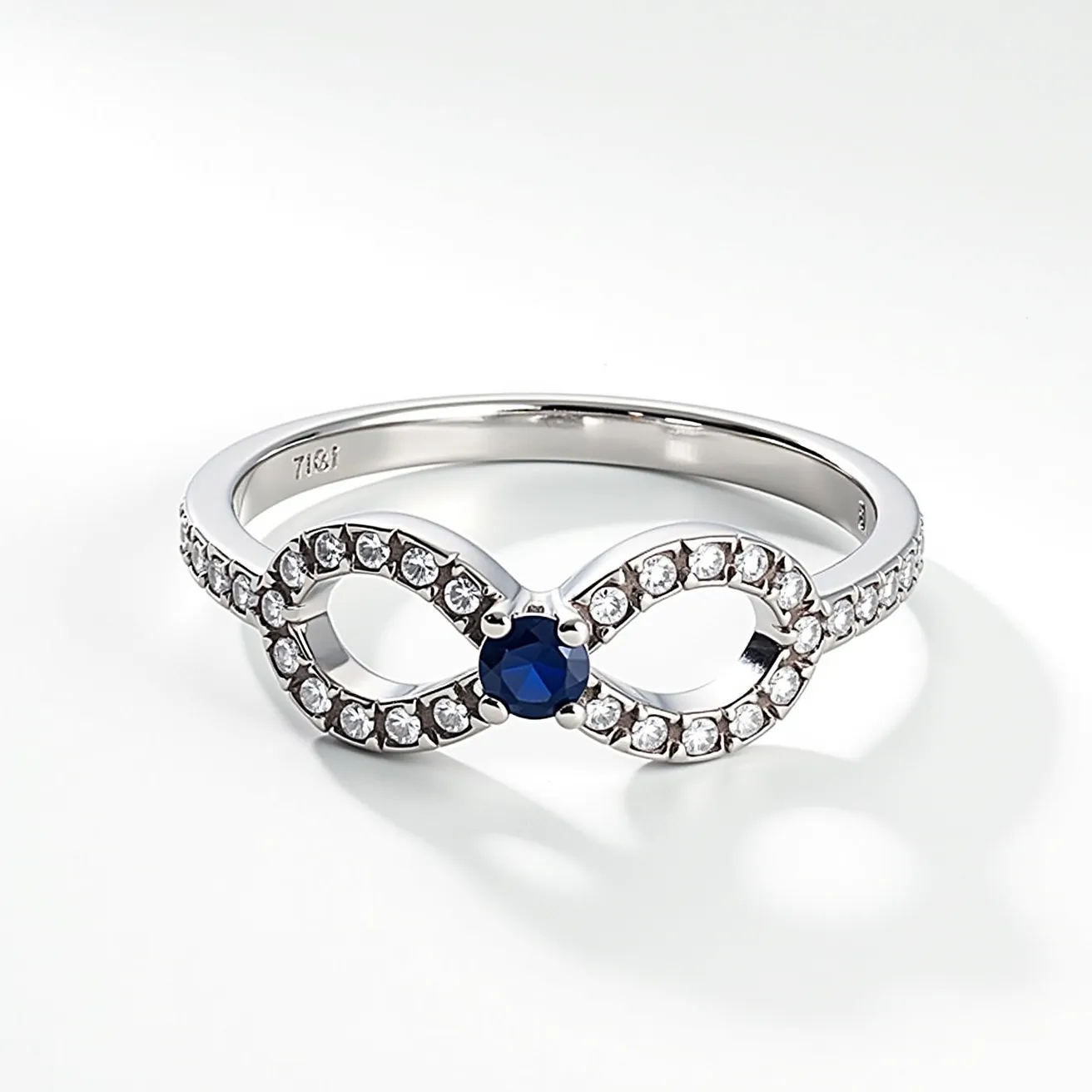 This two birthstone ring features a sleek metal band, likely crafted from silver or white gold, designed in an elegant infinity symbol. The centerpiece showcases two distinct gemstones; a deep blue sapphire cut in a round shape sits prominently within a secure prong setting, surrounded by a series of smaller, clear accent stones, presumably diamonds or cubic zirconia, which extend along the band. The arrangement offers a sophisticated, timeless appearance with a seamless integration of the stones into the ring's graceful design.