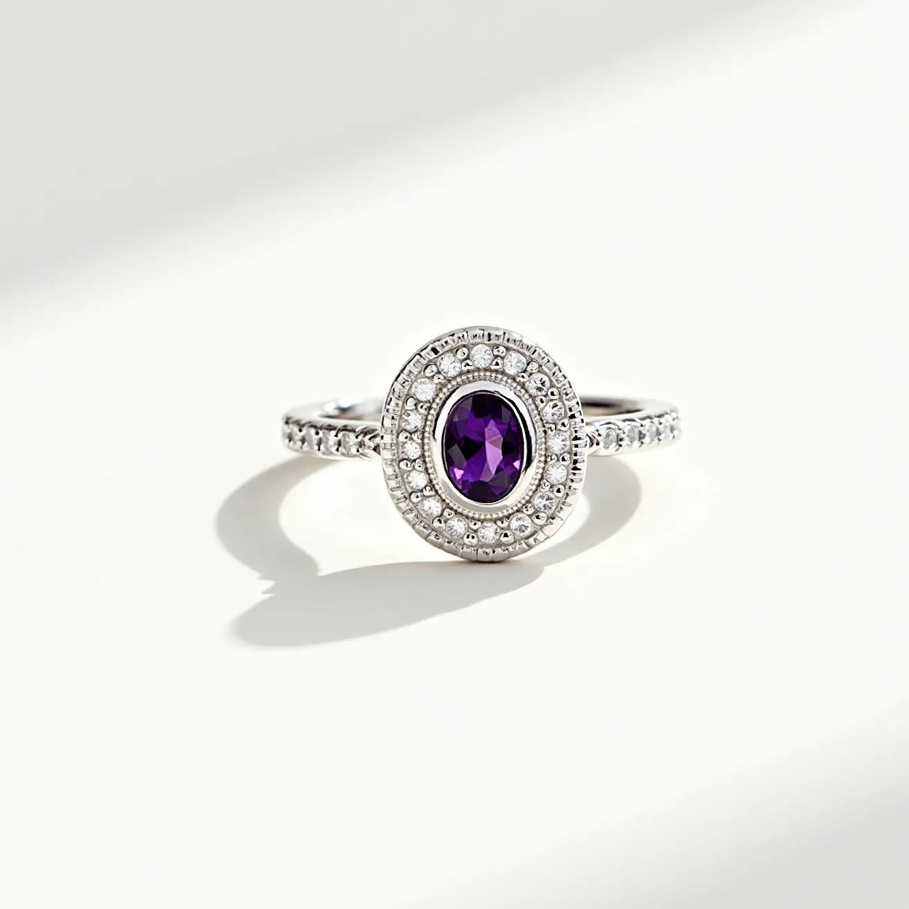 This two birthstone ring features a prominent oval-cut amethyst at its center, surrounded by a halo of smaller white diamonds, enhancing its brilliance and contrast. The band is crafted from a polished white metal, likely white gold or platinum, and is adorned with a series of pavé-set diamonds, adding an elegant sparkle to the piece. The amethyst is set in a bezel or prong setting, ensuring security while showcasing its rich purple hue. The ring is a seamless piece without any visible clasps, designed to slip effortlessly onto the finger.