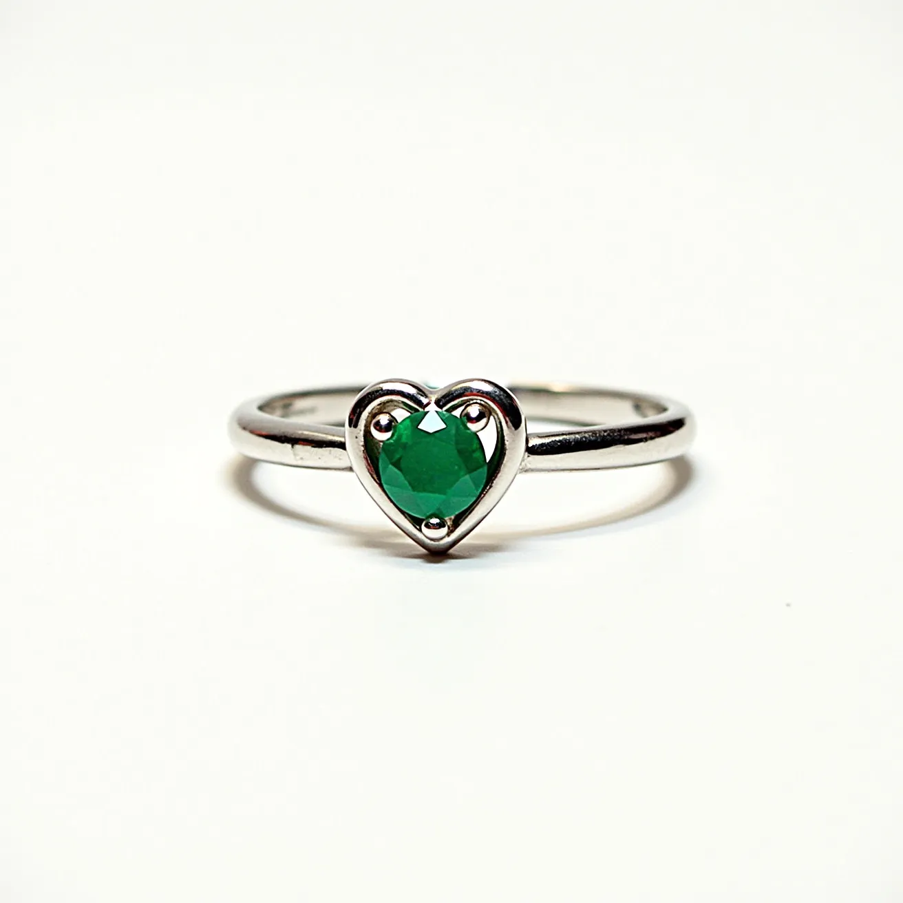 This two birthstone ring features a sleek, polished band, likely made from a silver-colored metal, such as sterling silver or white gold. It showcases a single, vivid green gemstone, possibly an emerald, cut in a round shape and prominently set within a heart-shaped setting. The gemstone is held securely by three prongs integrated into the design, emphasizing its central position on the band. The ring combines elegance with a touch of personalization, symbolized by the solitary birthstone.