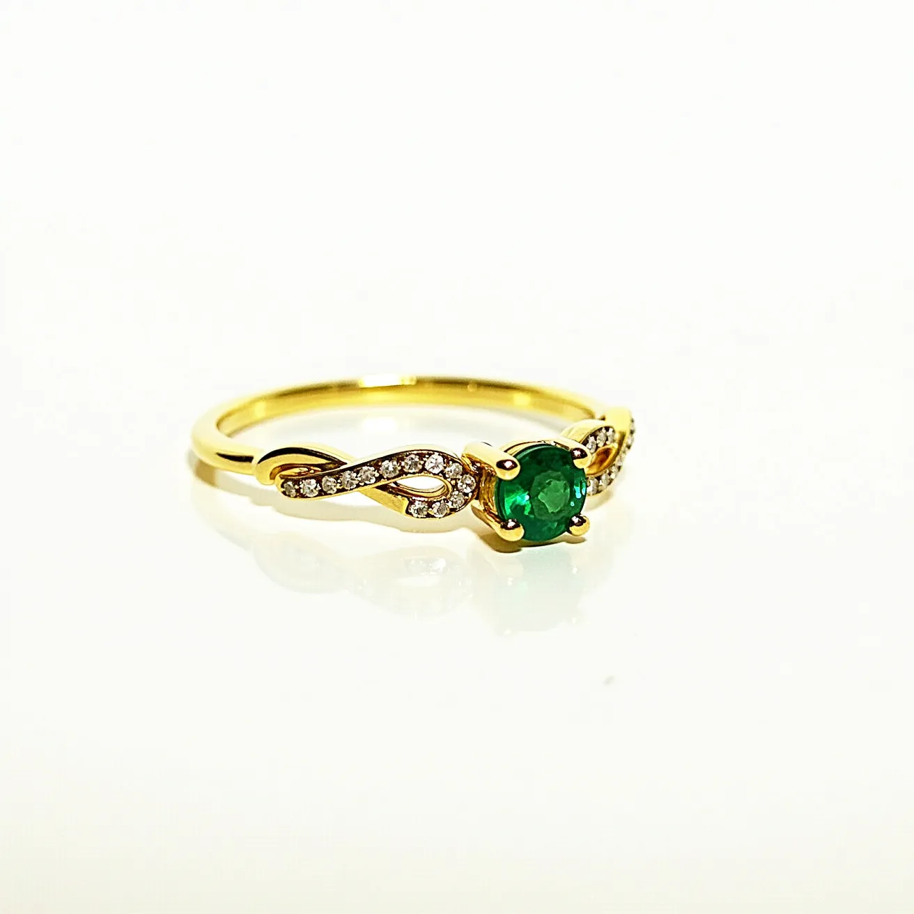 This vine engagement ring features a delicate gold band intricately designed in a vine pattern, adorned with small round diamonds set along the swirling vine in a pavé setting. At the center of the design lies a captivating round emerald, held securely in place by a classic four-prong setting, showcasing its vibrant green hue. The combination of the rich gold with the sparkling diamonds and the deep green emerald creates a harmonious and elegant appearance, with the vine motif adding a touch of natural beauty.