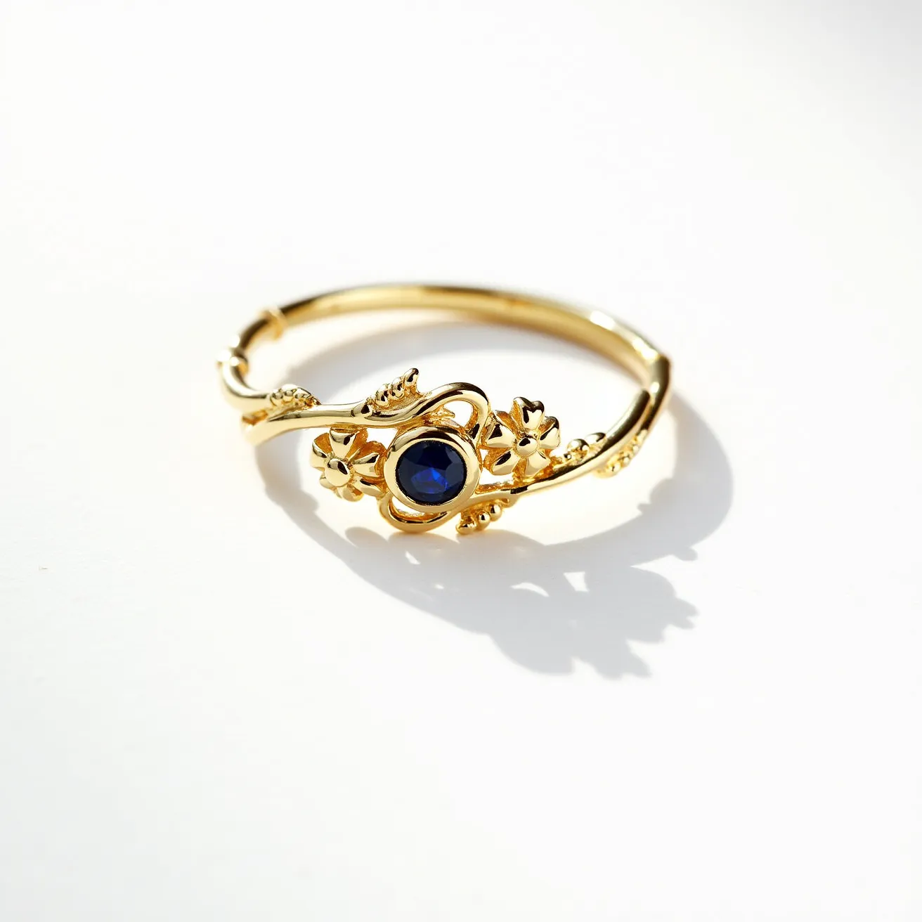 This vine ring is crafted from a lustrous gold material, featuring a beautifully intricate design reminiscent of delicate vines adorned with small flowers. At its center, a striking round-cut blue gemstone is set in a secure bezel setting, enhancing its allure and elegance. The vine motif wraps gracefully around the band, creating an ornate and nature-inspired appearance. The craftsmanship highlights a seamless integration of the elements, making it a captivating piece of jewelry.