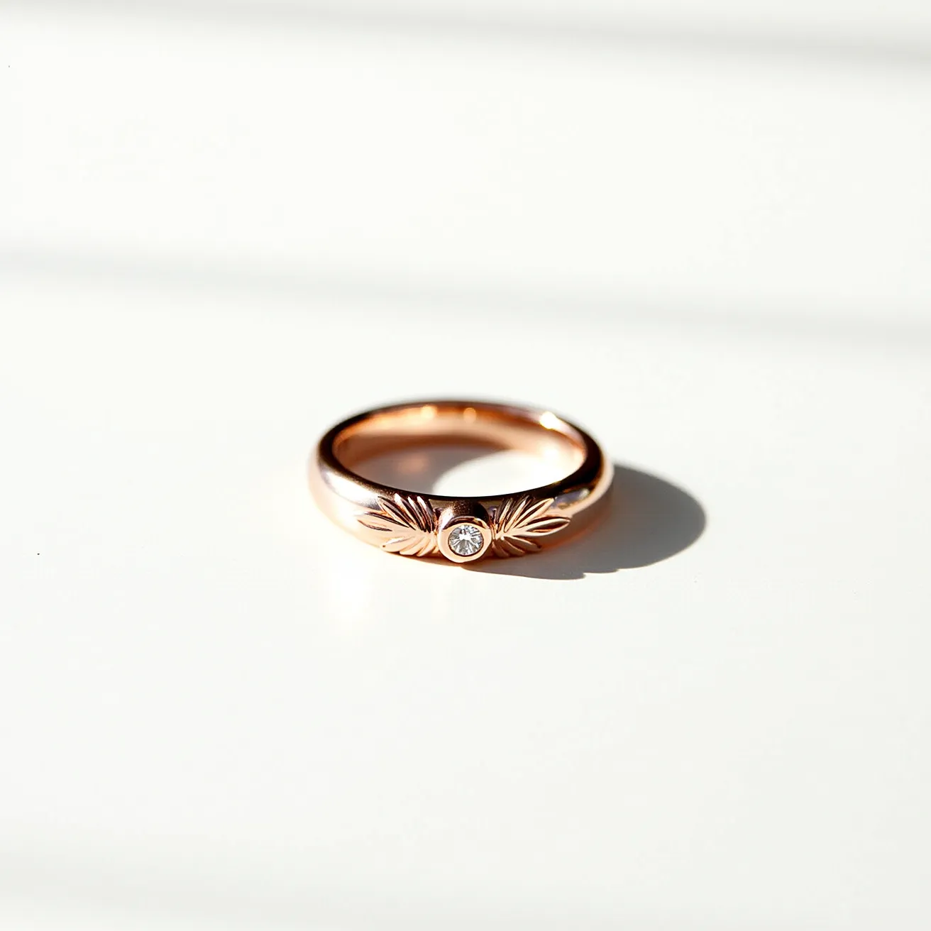 This wedding band is crafted from rose gold, featuring an elegant and delicate design. It showcases a round-cut diamond centrally set in a bezel setting, enhancing the stone's brilliance while providing a secure hold. The band includes intricate engravings resembling leaves or feathers flanking the diamond, adding a touch of nature-inspired artistry to the piece. The smooth, polished finish of the rose gold enhances its warmth and complements the sparkle of the diamond, creating a beautiful and timeless look.