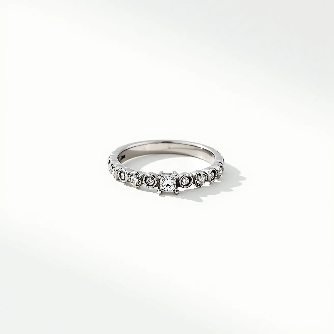 This wedding ring features a sleek metal band, likely platinum or white gold, adorned with a series of small round diamonds set in delicate bezel settings. The centerpiece of the ring is a square-cut diamond in a classic prong setting, adding a touch of elegance and sophistication. The round diamonds encircle the band partially, enhancing the ring's brilliance and providing a cohesive design. This combination of gemstones and settings creates a timeless and graceful appearance, ideal for a wedding band.