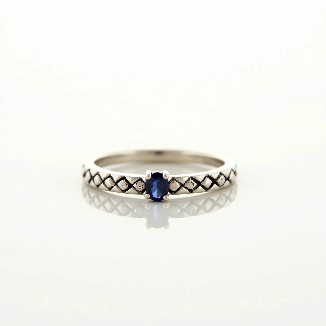 This wedding ring features a band crafted from what appears to be a polished silver material, adorned with a quilted pattern along its circumference. At the center, a dark blue gemstone, likely a sapphire, is set in a four-prong setting, adding a striking focal point to the ring. The gemstone is cut in an oval shape, enhancing its reflective qualities and depth of color. The overall design combines elements of elegance with a touch of intricate detailing, creating a unique and memorable piece.