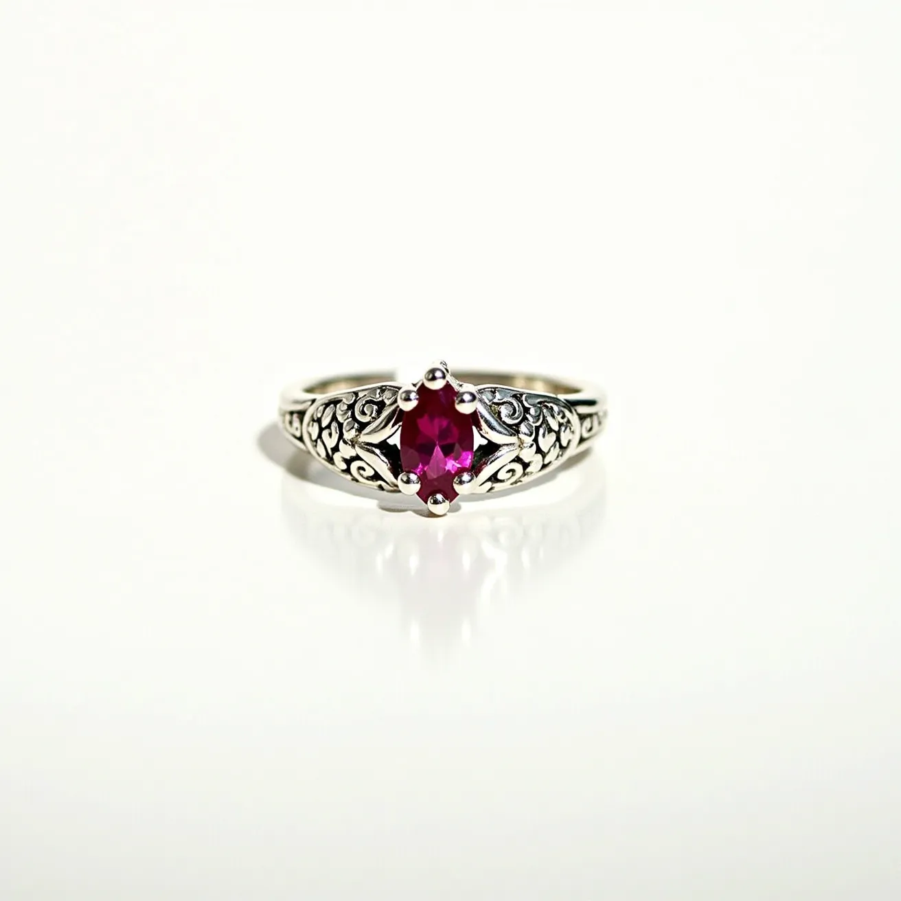 This wedding ring features a beautifully intricate design with a silver band that showcases detailed scrolling patterns. At its center, there is a vibrant oval-cut red gemstone, likely a ruby, securely held in place by a classic prong setting. The prongs are gracefully positioned to ensure both security and maximum exposure of the stone's brilliance. The design of the band, with its ornate engravings, adds to the ring's elegance and vintage aesthetic.