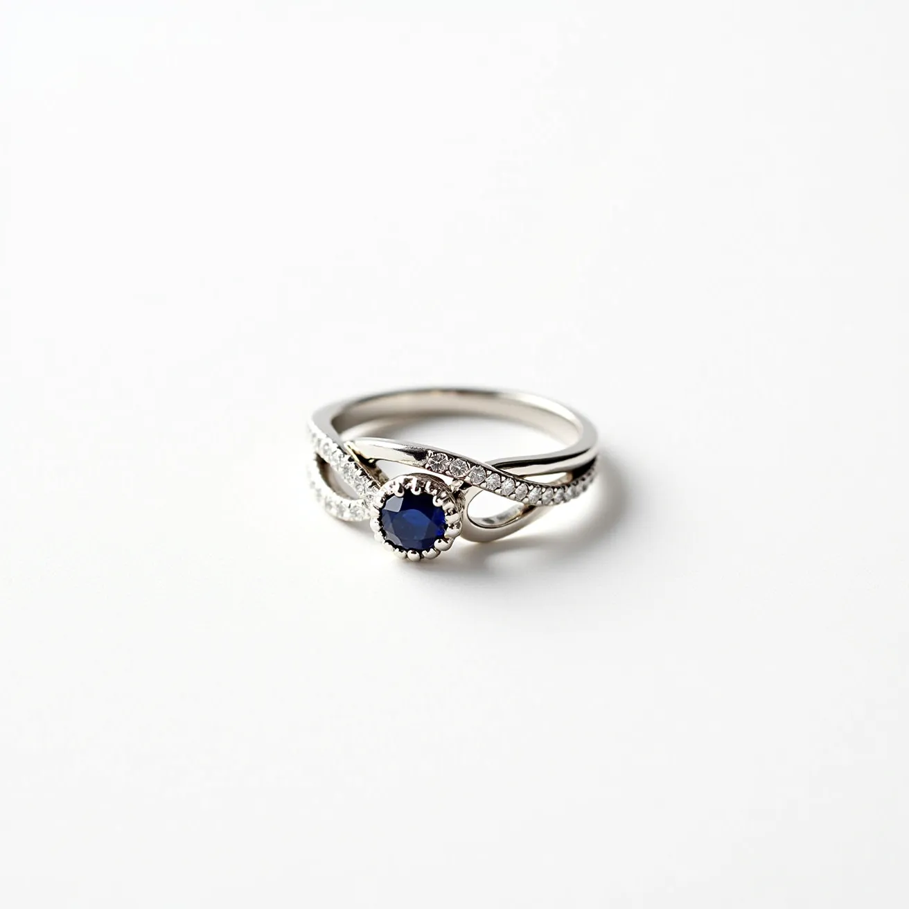 This wedding ring features a white metal band, likely platinum or white gold, intricately designed with an intertwining pattern. At its center is a round-cut sapphire, deep blue in color, held in place by a decorative prong setting that adds an elegant touch. Surrounding the central sapphire are multiple small, round-cut diamonds set into the band, enhancing its brilliance and sophistication. The design is seamless, with no visible clasp or attachment, emphasizing its continuous and harmonious form.