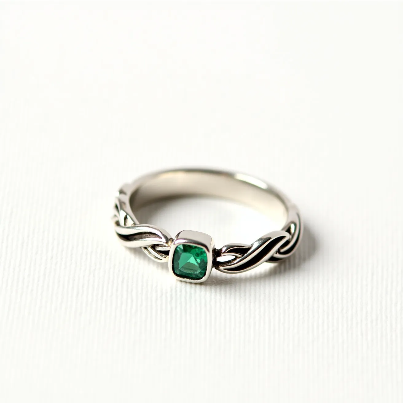 This wedding ring features a silver band with an intricate Celtic knot design. At the center of the ring is a square-cut green gemstone, likely an emerald, which is set in a bezel setting. The combination of the detailed knot pattern and the vibrant central stone gives the ring a unique and elegant appearance. The ring does not incorporate any clasps or attachments, maintaining a continuous and seamless design.