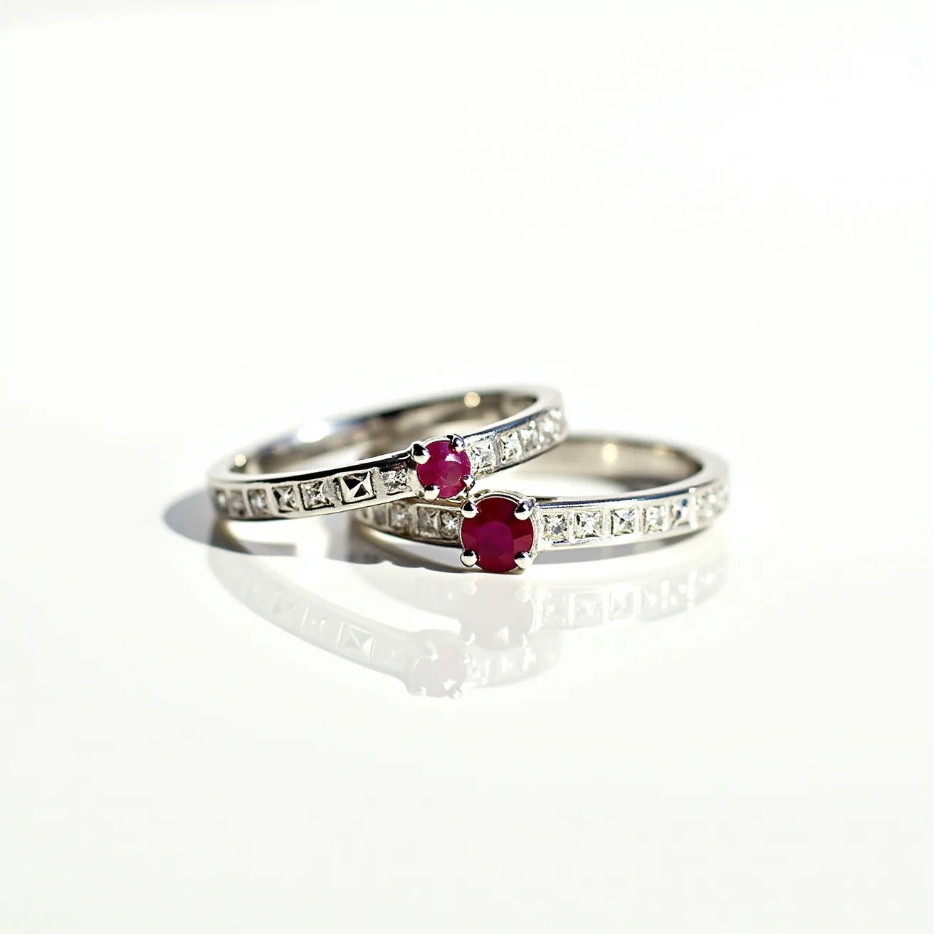 These wedding ring sets feature a stunning and elegant design crafted from what appears to be a silvery metal, possibly white gold or platinum. Each ring is adorned with a central round-cut ruby set in a classic four-prong setting, which adds a bright pop of color. Along the band, these rings are embellished with a series of small square-cut diamonds set in precise rows, enhancing their sparkle and overall sophistication. The combination of the vibrant red rubies and the shimmering diamonds creates an eye-catching contrast, perfect for celebrating a timeless union.