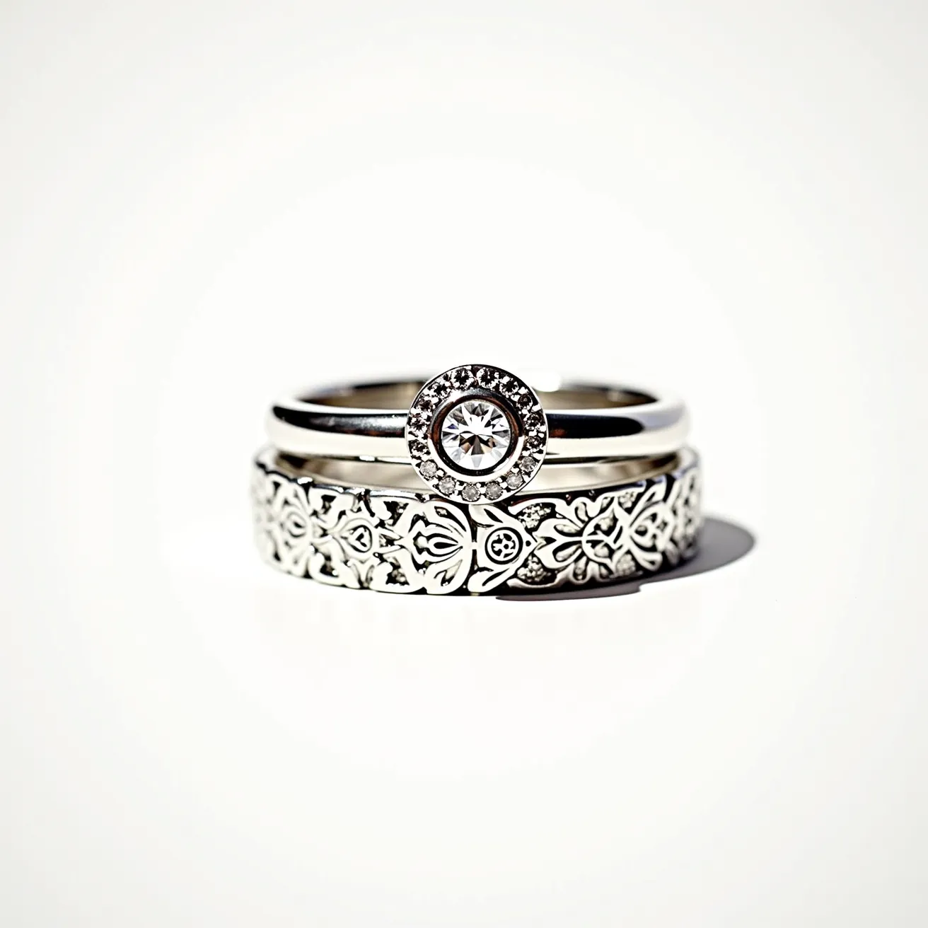 These wedding ring sets feature a combination of sleek metal bands and exquisite detailing. The top ring showcases a prominent round-cut gemstone, set in a halo style with smaller stones encircling it, creating a brilliant focal point. The band appears to be made from a lustrous silvery metal, giving it a classic and elegant look. The accompanying band displays intricate floral motifs etched into the surface, adding a touch of artistic flair and texture to the ensemble. Together, these rings combine timeless elegance with delicate artistry, making them a striking set for a wedding or engagement.