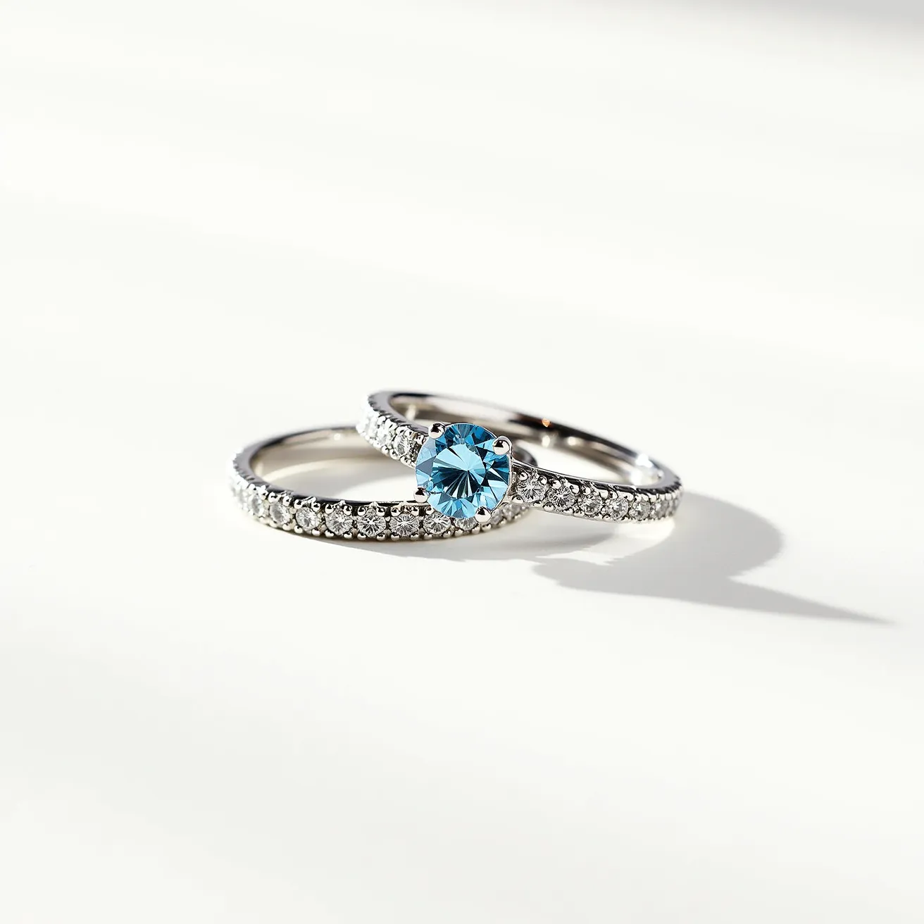 These wedding ring sets feature a beautiful combination of sleek, polished metal bands adorned with a series of small, sparkling white stones set around the circumference, likely diamonds or cubic zirconia, arranged in a pavé setting. The highlight of the set is the engagement ring, which showcases a striking round-cut blue gemstone in a prong setting, offering a vibrant contrast to the surrounding sparkles. The matching wedding band complements the engagement ring, featuring a uniform row of the same smaller stones but without a central gemstone, creating a harmonious and elegant duo.