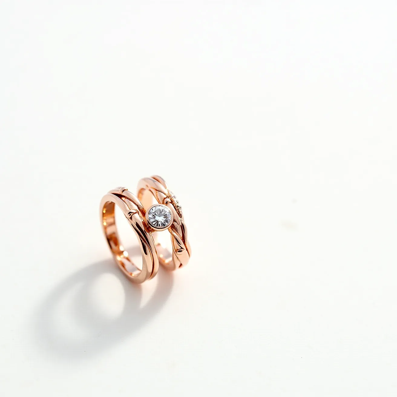 These wedding ring sets feature a warm rose gold material, giving them a romantic and elegant appearance. The set includes a striking central ring adorned with a prominent round-cut gemstone, likely a diamond, set in a bezel setting that enhances its brilliance and secures it in place. The accompanying band complements the main ring with a similar rose gold design and appears to have subtle detailing that matches the elegant theme of the set. The craftsmanship shows intricate attention to design, resulting in a harmonious and unified aesthetic between the two rings.