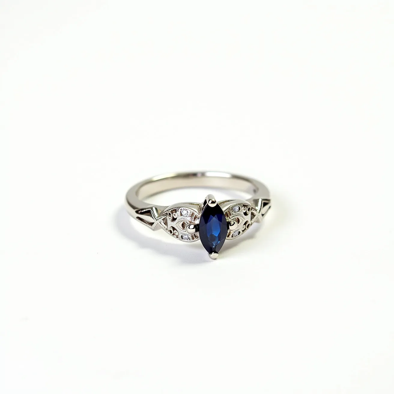 This western wedding ring is crafted from a polished metal, likely white gold or platinum, and features an elegant design with a marquise-cut sapphire centerpiece. The sapphire is secured by a prong setting, adding both stability and style to the ring. On either side of the central stone are intricate metalwork designs, possibly with small accent diamonds or similar stones set within the pattern, enhancing the ring's sophistication. The band itself is smooth and rounded, offering comfort and a timeless look.