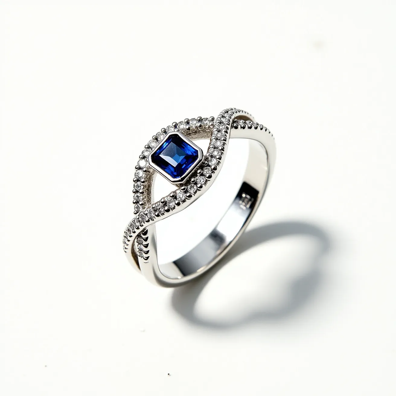 This western wedding ring features an elegant design crafted from a silvery metal, likely white gold or platinum, known for its durability and classic appeal. The centerpiece is a striking blue square-cut gemstone, possibly a sapphire, set securely in a prong setting. Surrounding the central stone is a series of small, clear, round-cut diamonds embedded in a pavé setting, which add a sparkle and enhance the brilliance of the central sapphire. The band displays a unique twist design, complementing the ensemble and adding a touch of sophistication to the overall piece.