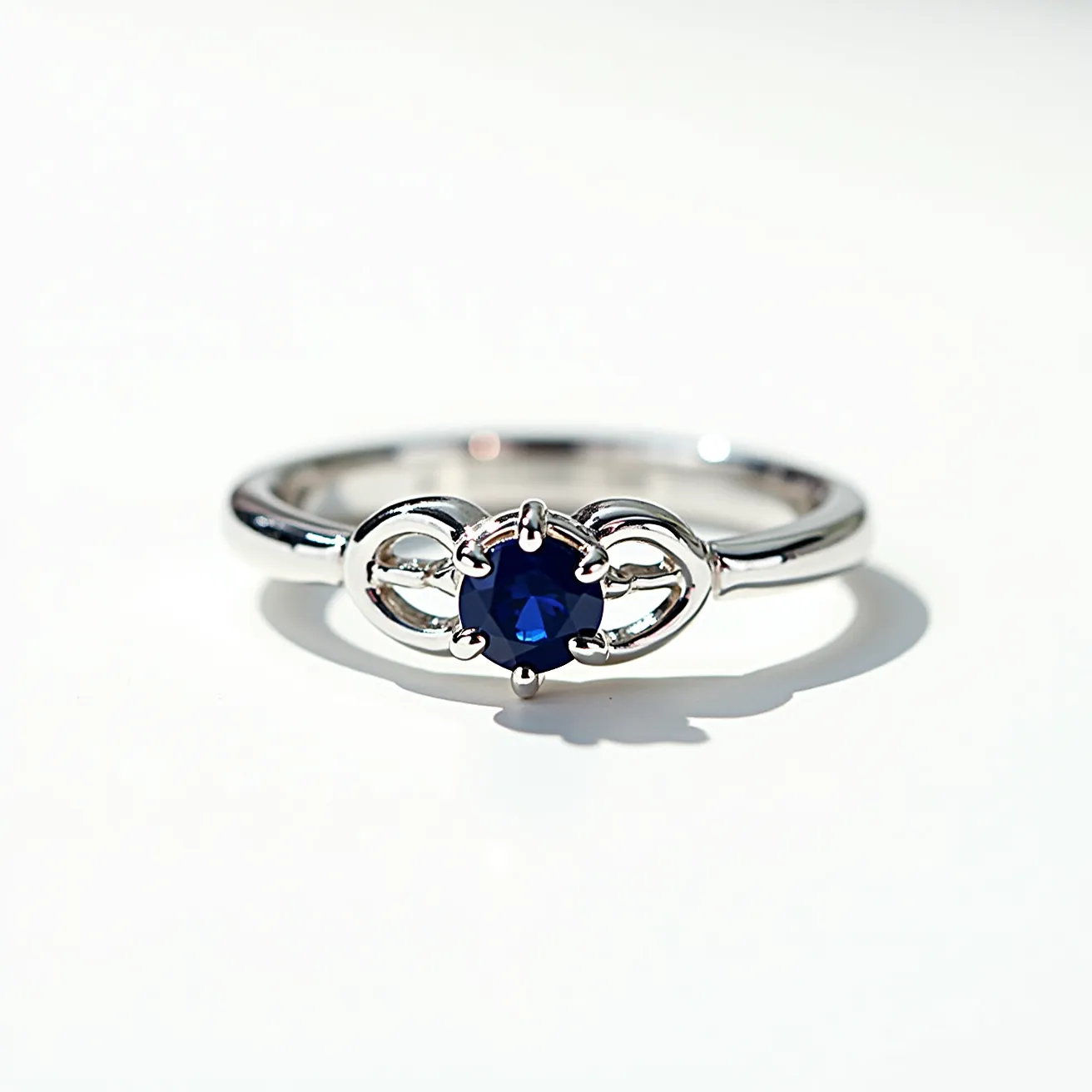 This white gold ring features a central blue gemstone, possibly a sapphire, cut in a round shape and secured within a prong setting. The elegant design incorporates intricate metalwork on either side of the gemstone, adding a touch of sophistication to the overall appearance. The ring’s band is smoothly polished, contributing to its refined and timeless aesthetic.