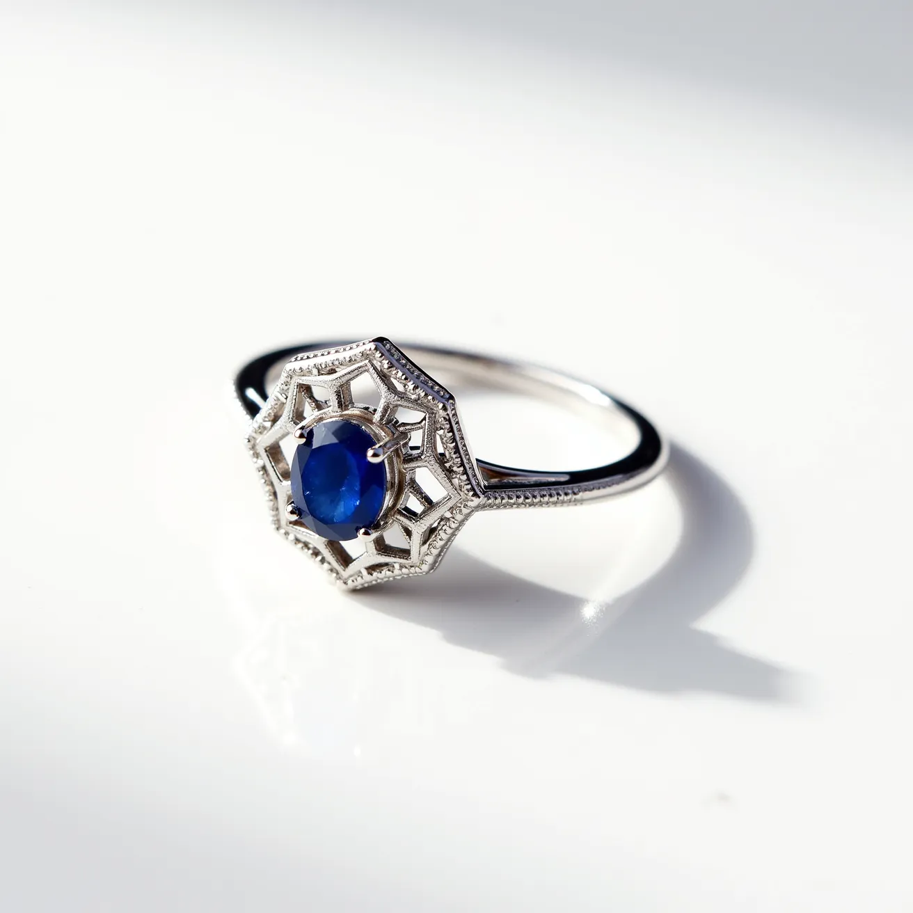 This white gold ring features an intricately crafted setting that holds a prominent blue gemstone, likely a sapphire, with an oval cut. The gemstone is securely set within a hexagonal, open-work design, bordered by delicate milgrain detailing that enhances the ring’s elegance. The prong setting ensures that the sapphire is prominently displayed, capturing light beautifully. The band is crafted from polished white gold, offering a sleek and sophisticated finish, with no visible clasps or additional attachments.