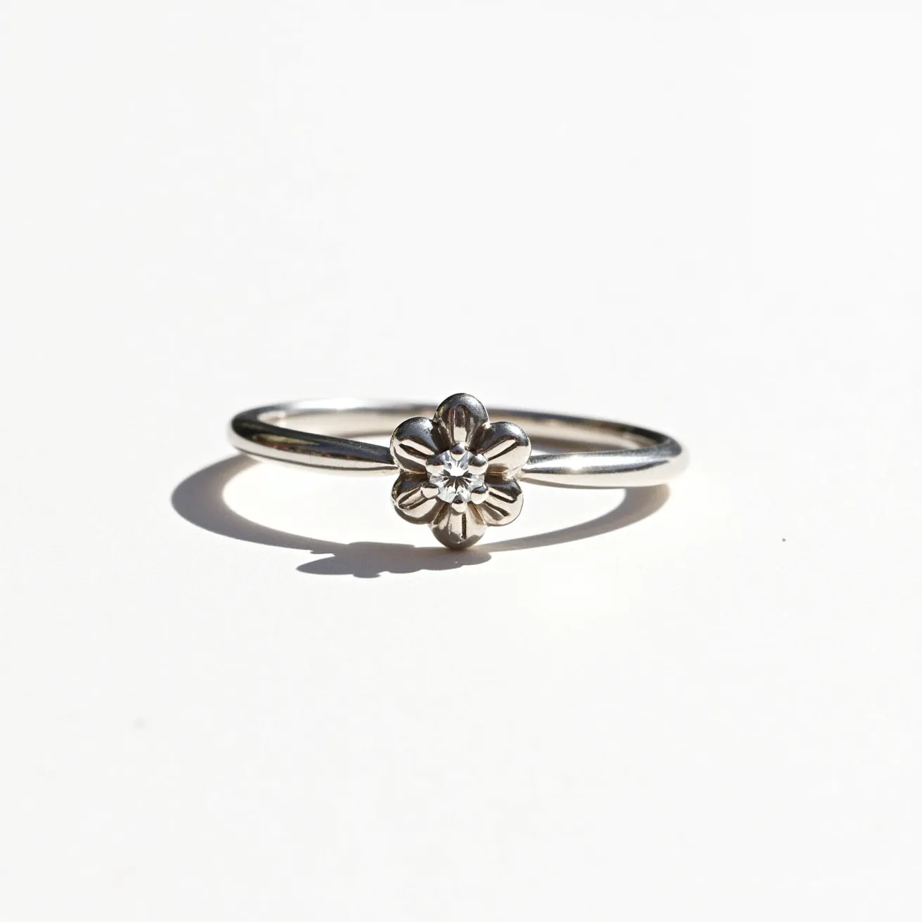 This white gold ring features a delicate floral design at its center, with a small diamond or crystal set in the middle. The gemstone appears to be round cut, adding a subtle sparkle to the piece. The setting is integrated seamlessly with the petal-like design, creating an elegant and unified look. The band is slim and polished, complementing the simplicity of the flower motif. There are no additional clasps or attachments, highlighting the minimalist and refined aesthetic of the ring.